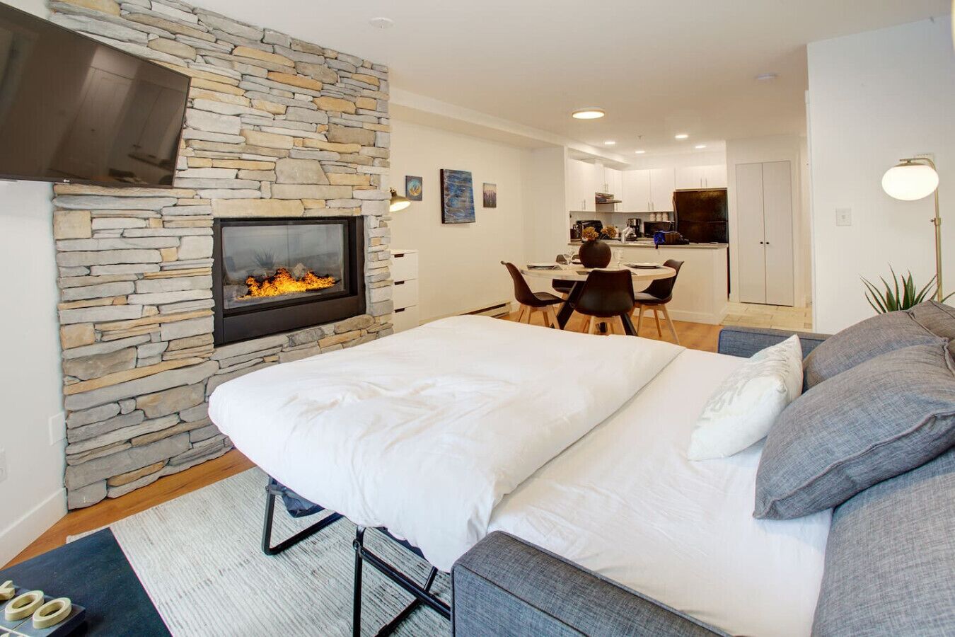 A bedroom with a stone fireplace, a bed with grey and white bedding, a grey sofa, and a dining area in the background.