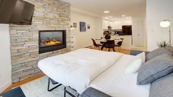 A bedroom with a stone fireplace, a bed with grey and white bedding, a grey sofa, and a dining area in the background.