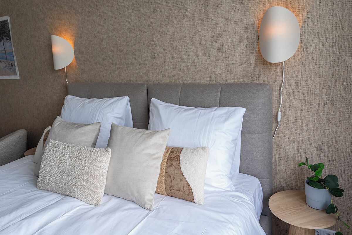 Una cama bien hecha con ropa de cama blanca y varias almohadas, una mesita de madera redonda con una planta, y dos lámparas fijadas a la pared.
