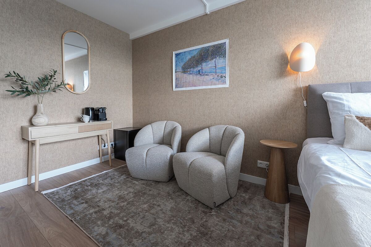 Een moderne hotelkamer met een bed, twee fauteuils, een nachtkastje, een klein bijzettafeltje, een spiegel, een schilderij en een wandlamp.