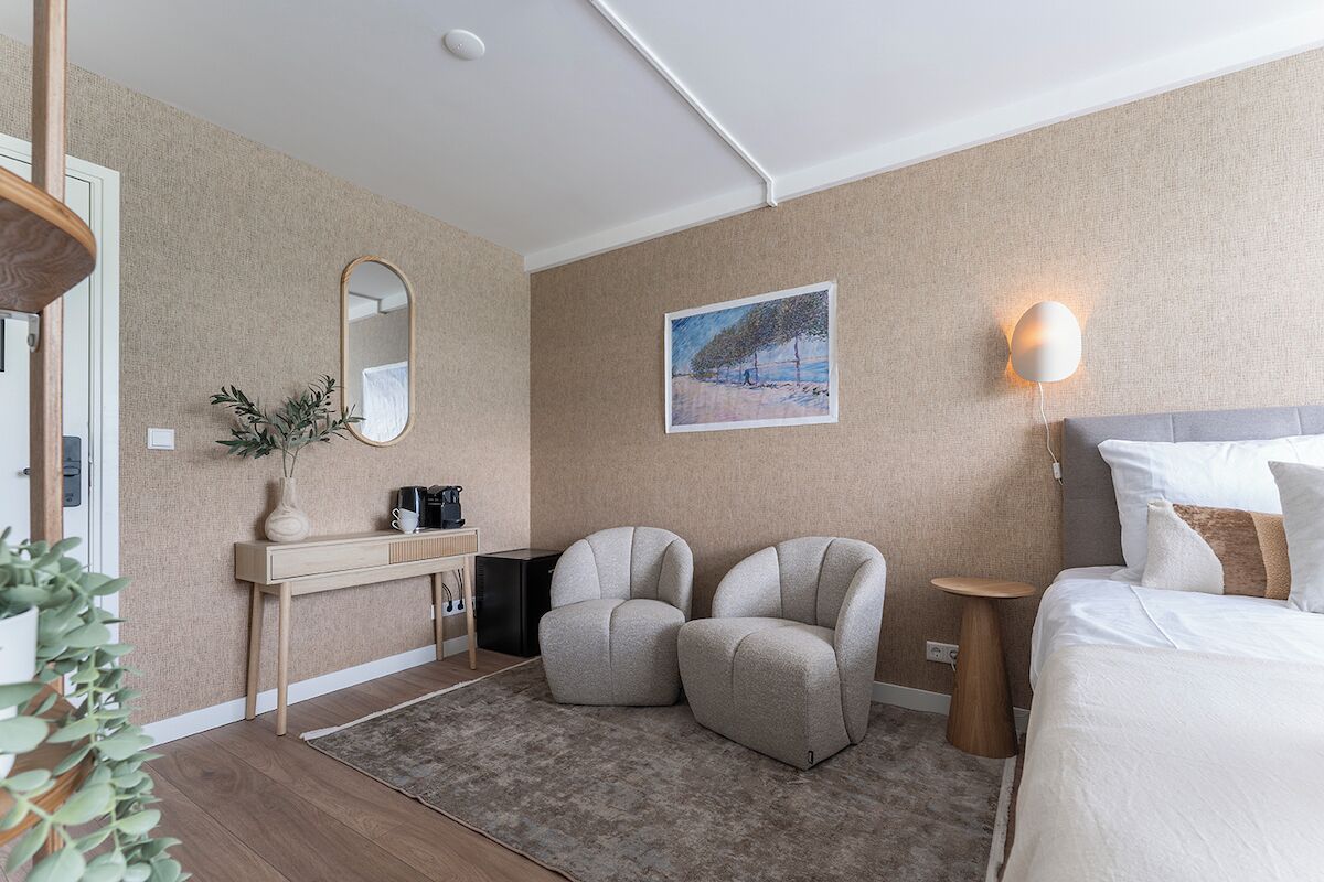 Una habitación de hotel moderna con una cama, dos sillones, una mesita, un espejo y un cuadro en la pared.