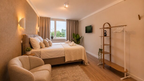 Un dormitorio con cama, una silla, televisión, ventana con cortinas y un perchero de madera.