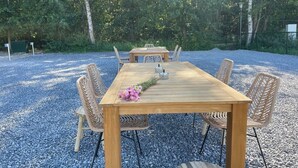Une table en bois, entourée de quatre chaises en osier, posée sur du gravier, avec des arbres et une clôture en arrière-plan.