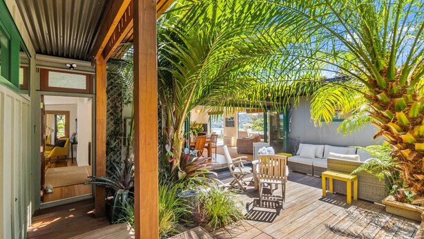 A wooden deck with outdoor furniture, a white sofa, and a large palm tree. - The Zen House - Our Island (Waiheke Island)