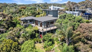 A modern house with a large deck, surrounded by dense vegetation and hills. - The Zen House - Our Island (Waiheke Island)