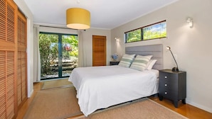 A bedroom with a bed, a nightstand, a lamp, a window, and a door leading to a balcony. - The Zen House - Our Island (Waiheke Island)