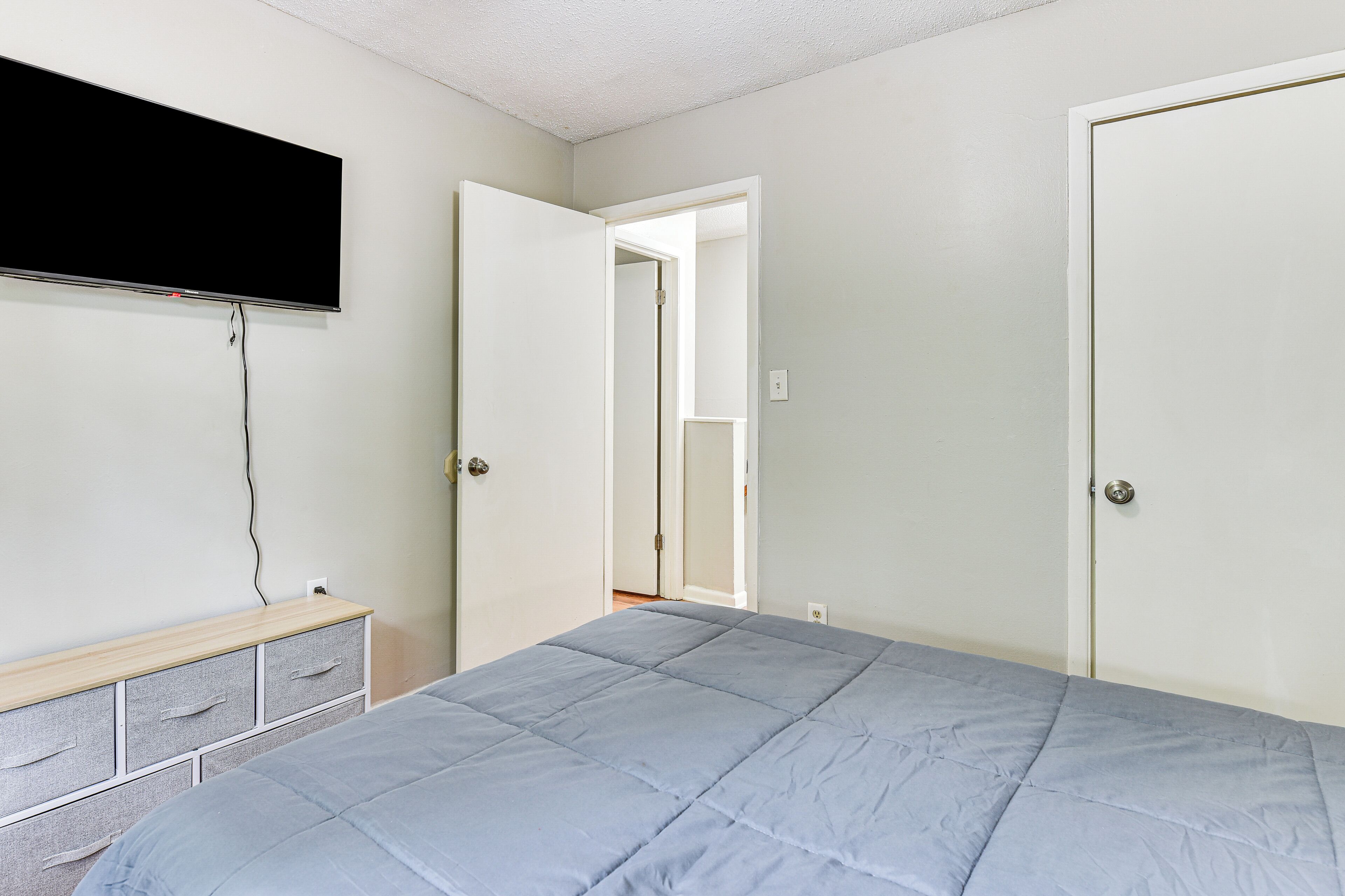 Ein Schlafzimmer mit einem Bett, einem an der Wand befestigten Fernseher, einer Kommode und zwei Türen zu anderen Räumen.