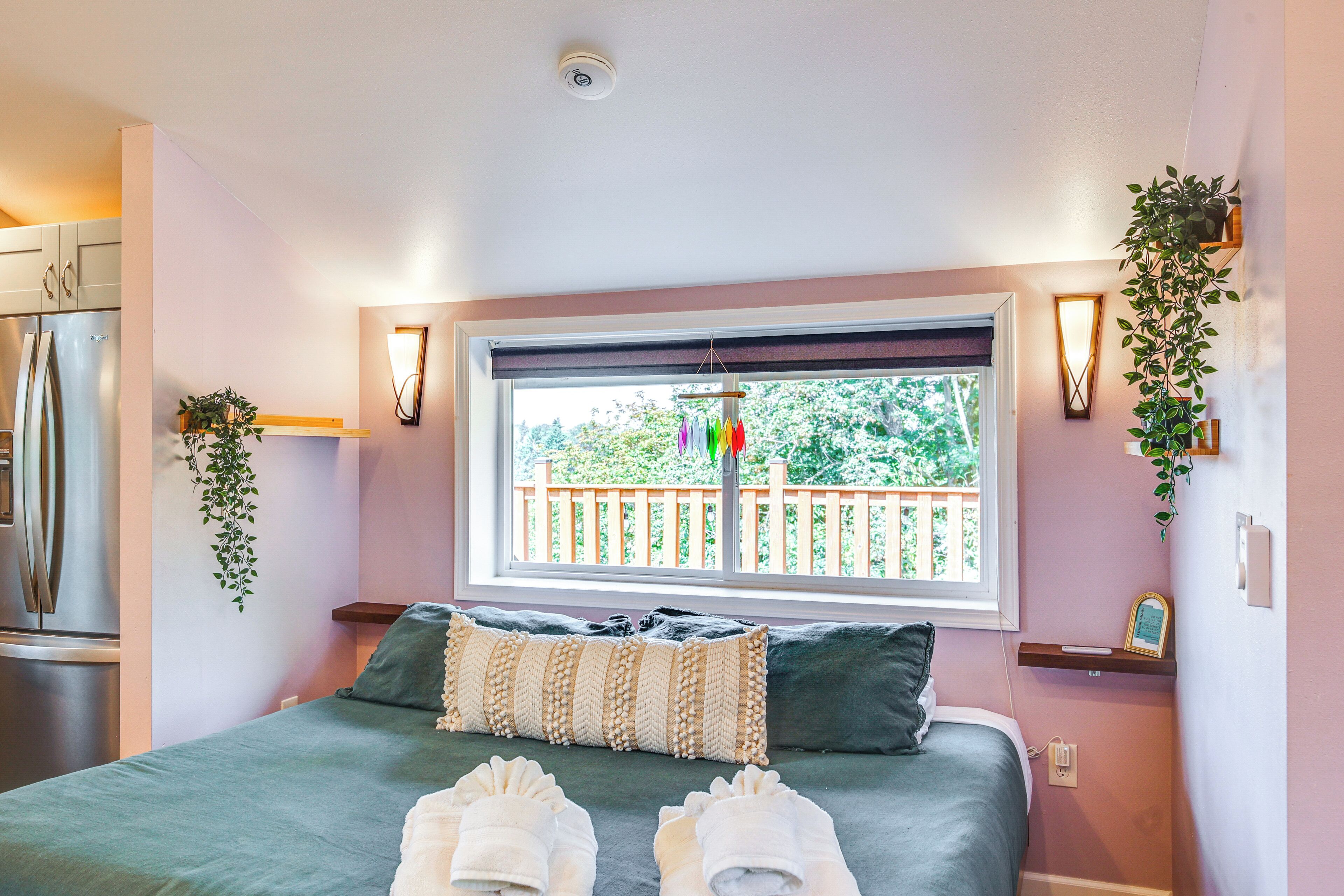 Een slaapkamer met een bed, kussens, een raam met uitzicht op een hek, een plant en een wandlamp.