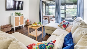 A living room with a sofa, a television, a table with fruits and a plant, and a view through large windows. - 'wandering Waves!' w/ Private Beach & Pool Access (St. Augustine)