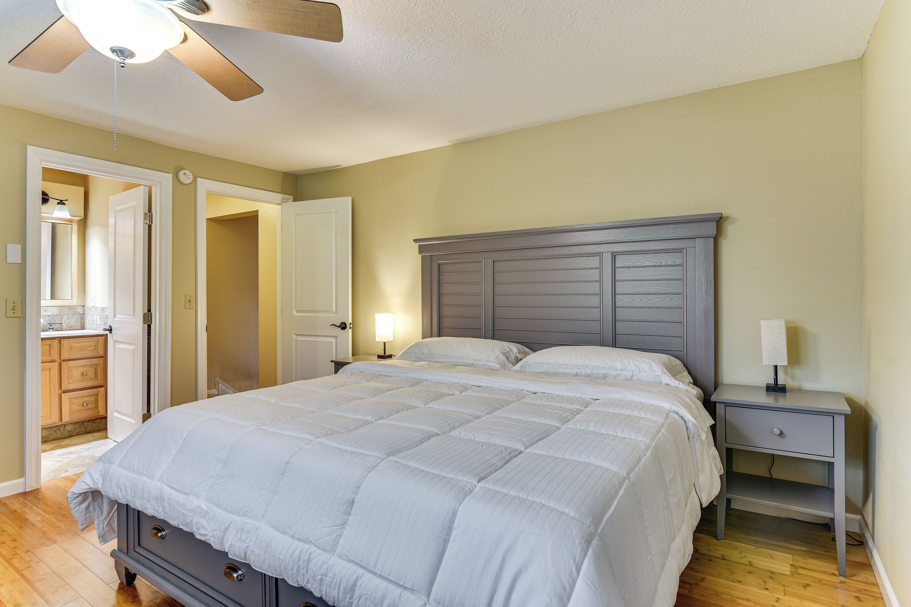 A bedroom with a large bed, a nightstand, a ceiling fan, and a door leading to a bathroom.