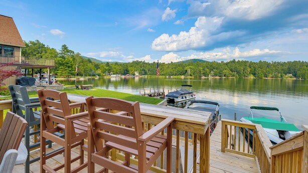Terraza de madera con sillas y una mesa con vistas a un lago, con barcos amarrados y una montaña al fondo.