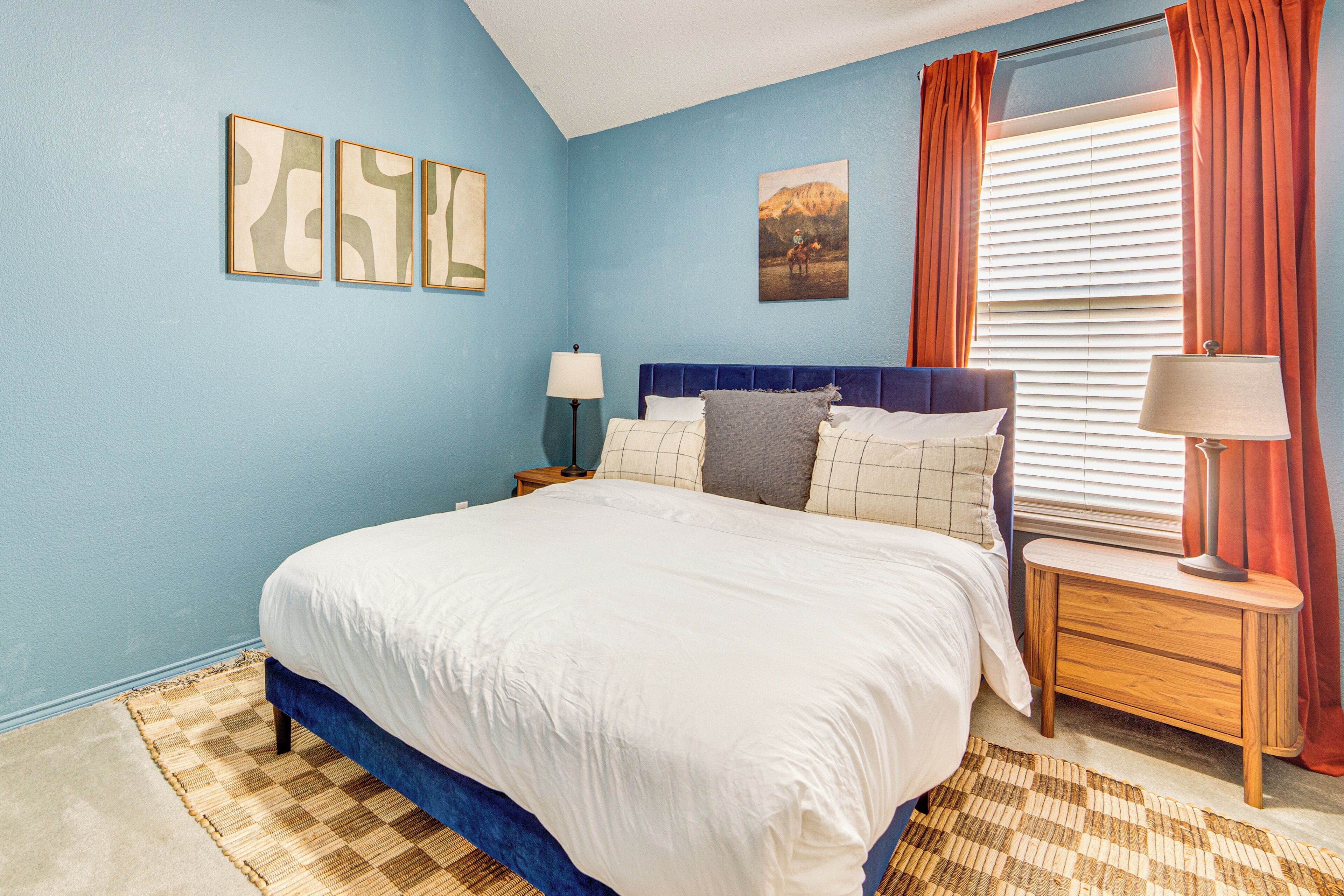 Une chambre à coucher avec un grand lit, deux tables de chevet, une fenêtre avec des rideaux et des œuvres d’art murales.