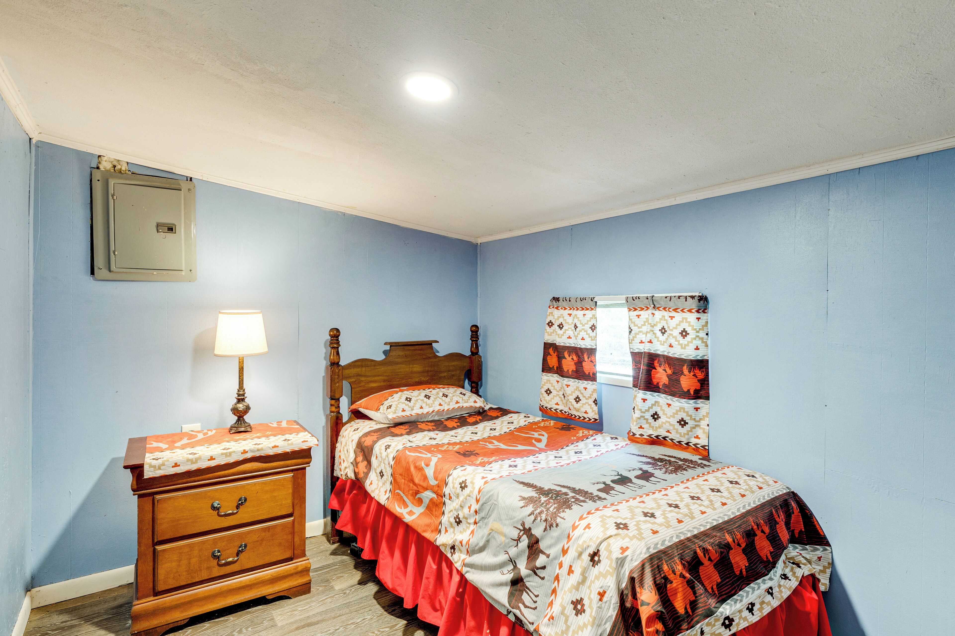 A bedroom with a bed, a nightstand, a lamp, and a wall-mounted electrical box.