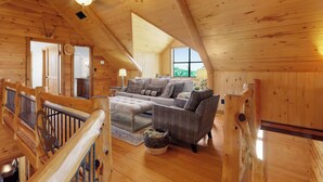 Un accogliente interno di una baita di legno con camera soppalcata, zona living con divano e poggiapiedi, e sala da pranzo.