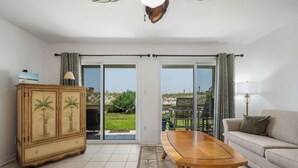 Un salón con sofá, mesa de madera, aparador con motivos de palmeras y vistas a un área exterior a través de un ventanal.