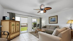 Una sala de estar con sofá, un mueble de madera con diseños de palmeras, un ventilador de techo y vistas al exterior a través de un ventanal.