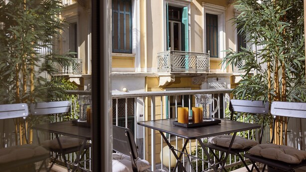 Een terras met tafels en stoelen, een balkon met een hekwerk, en een gebouw met ramen en luiken.