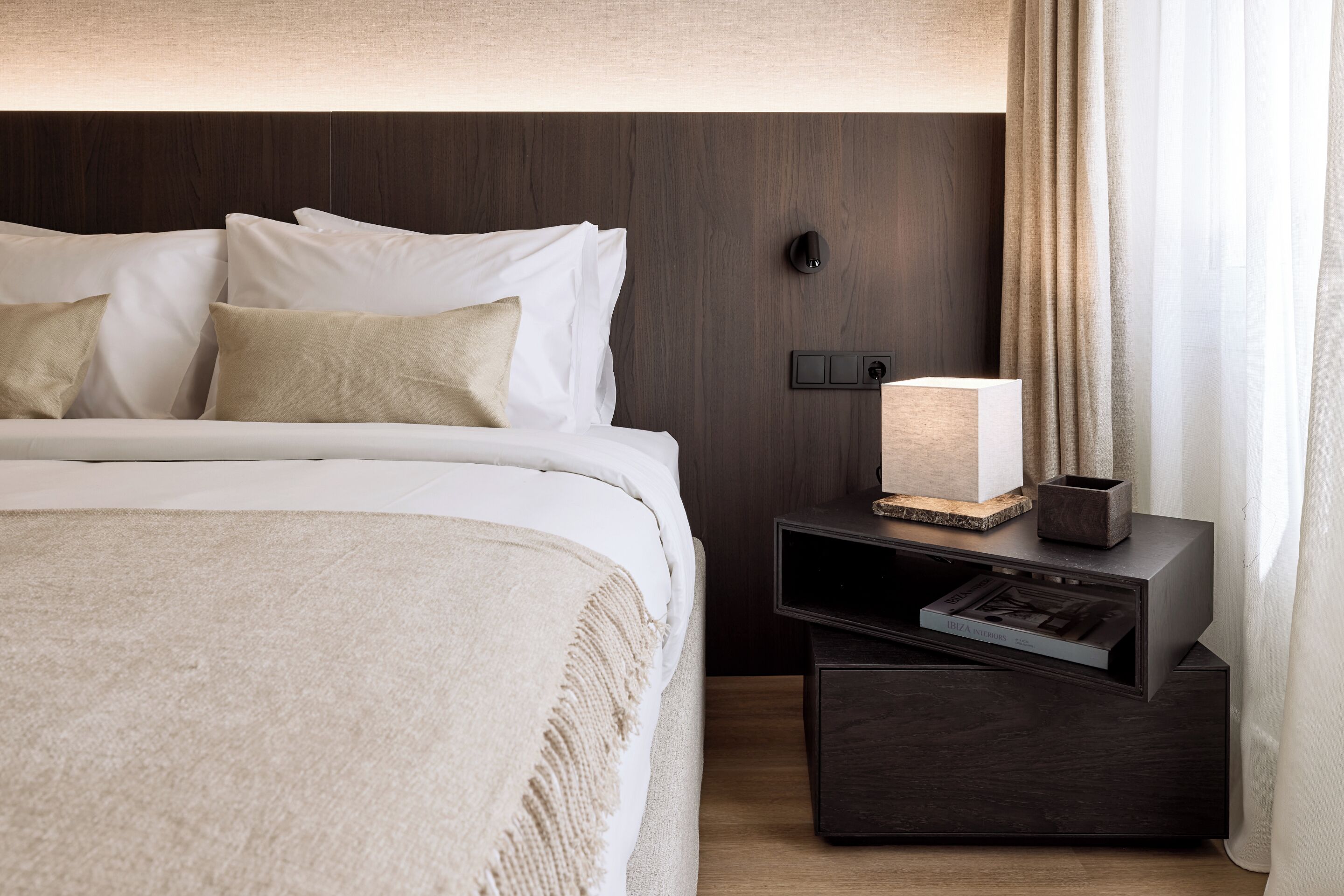 Una cama bien tendida con ropa de cama blanca y beige, cabecera de madera oscura, mesita de noche con lámpara y un pequeño adorno.