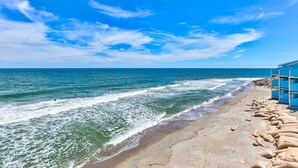 Una playa con olas, cielo despejado y un edificio azul con varias habitaciones.