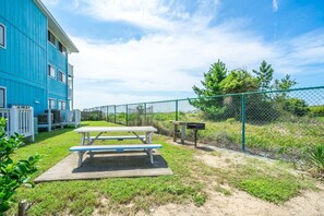 A picnic table in a grassy area with a chain-link fence and a blue building in the background.