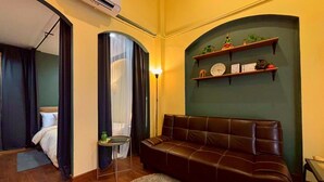 A modern hotel room with a leather sofa, a small table, a mirror, a bed, and a wall-mounted shelf with decorative items.