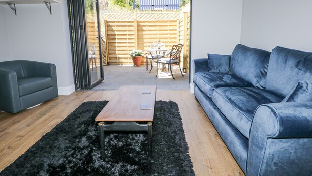 A living room with a blue sofa, a grey sofa, a wooden coffee table, and a black furry rug. There is a sliding glass door leading to a patio with a wooden fence, a table, and chairs. - Sunnyfield Cottage (Coulsdon)