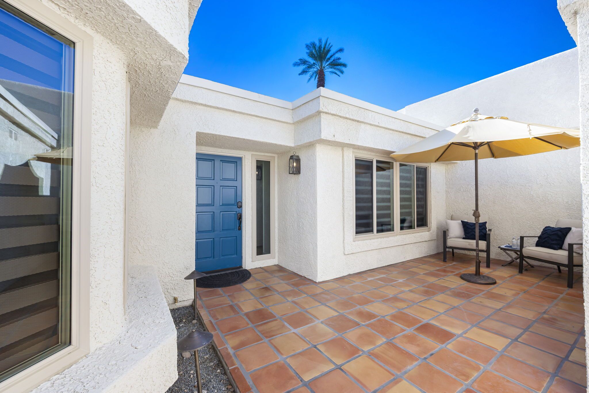 A white building with a blue door, a patio with tiled flooring, and a palm tree in the background.