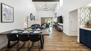 Un espace de salle à manger moderne avec une longue table, des chaises noires et un lustre. On aperçoit un canapé et un téléviseur au fond.