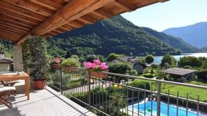 Terraza de madera con piscina, rodeada de plantas en macetas y vista a un lago y montañas.