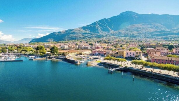 Un pueblo costero con un puerto deportivo, edificios a lo largo del malecón y montañas al fondo.