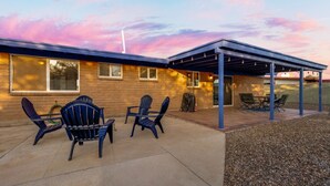A covered patio area with Adirondack chairs and a table, a brick building with large windows, and a clear sky.
