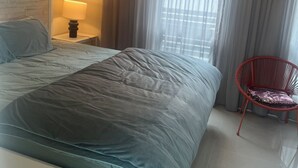 Une chambre à coucher moderne avec un grand lit, une table de chevet avec une lampe, un fauteuil rouge et des rideaux légers.