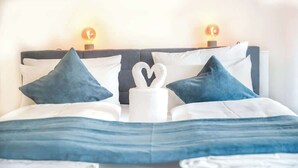 A neatly made bed with a heart-shaped towel and blue pillows, a dark headboard, and two wall-mounted lamps. - JH Apartments Ingolstadt | Penthouse Altstadt (Ingolstadt)