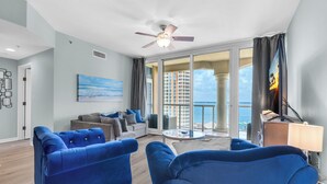 A modern living room with blue sofas, a ceiling fan, and a balcony with a view of the city.