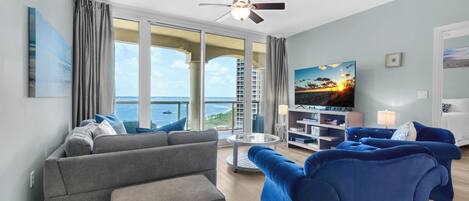 Un salon moderne avec un canapé gris, un canapé bleu et un téléviseur posé sur un meuble. Il y a un balcon avec vue sur l’océan.