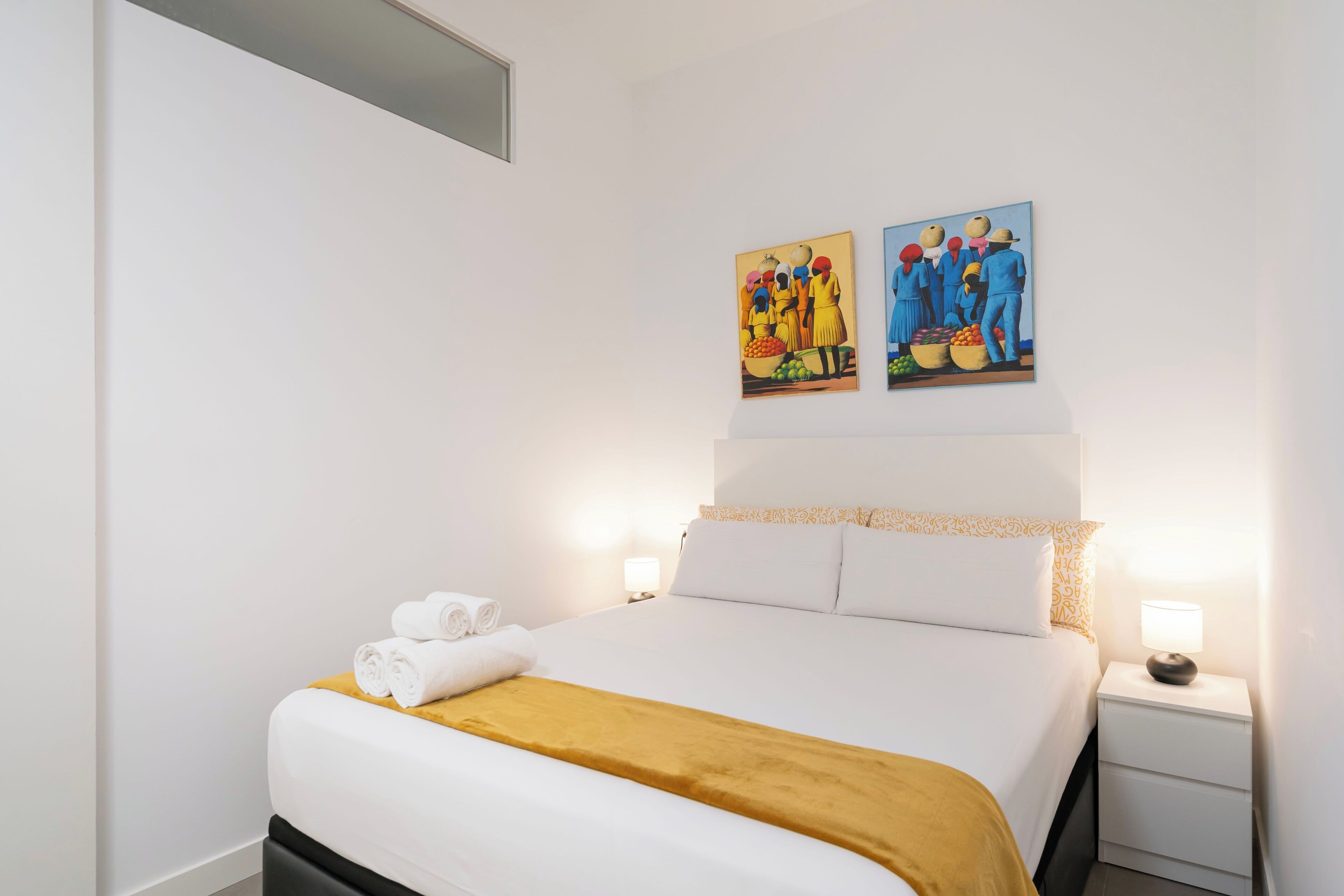 Una cama bien hecha con una colcha blanca y amarilla, dos cuadros en la pared y una mesita de noche con una lámpara.