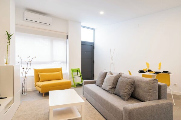 A modern living room with a sofa, a coffee table, a dining table with chairs, and a window with blinds. - Dobo Madrid Cochabamba (Madrid)