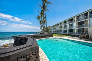 A hotel with a pool overlooking the ocean, featuring a stone wall and lounge chairs. - Kona Magic Sands#313:Top Floor,Direct Beachfront, Rarely Available Lanai unit (Kailua Kona)