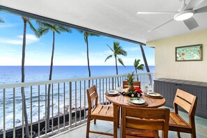 A balcony with a wooden table set for two, overlooking a beach with palm trees and the ocean. - Kona Magic Sands#313:Top Floor,Direct Beachfront, Rarely Available Lanai unit (Kailua Kona)