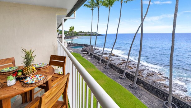 Un balcón con mesa y sillas de madera, con vistas a una playa con palmeras y una piscina.