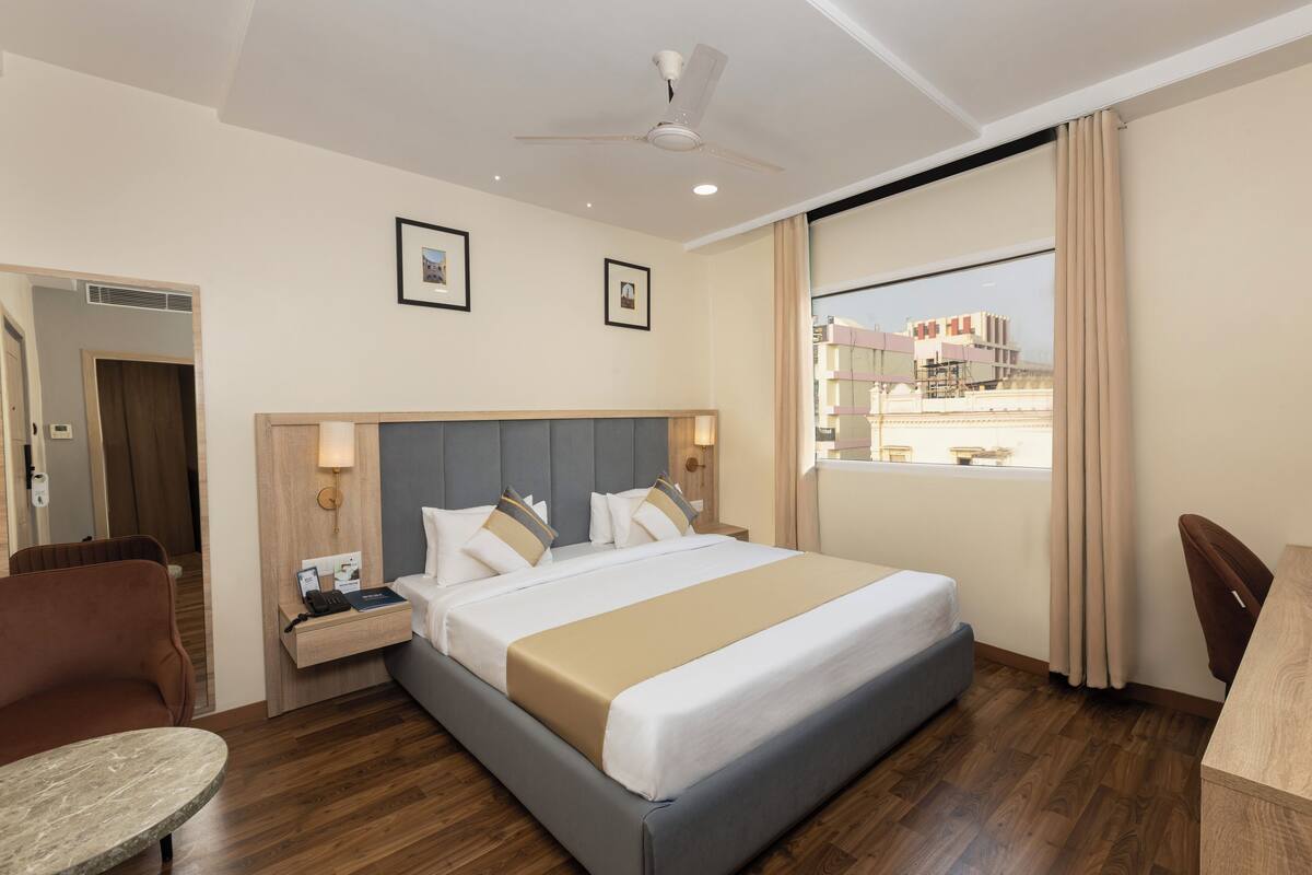 A hotel room with a bed, a desk, a chair, a table, a ceiling fan, and a window with curtains.
