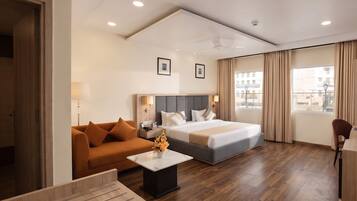 A hotel room with a bed, a sofa, a small table, and a desk. There are two windows with curtains, a ceiling light, and a wall-mounted picture.
