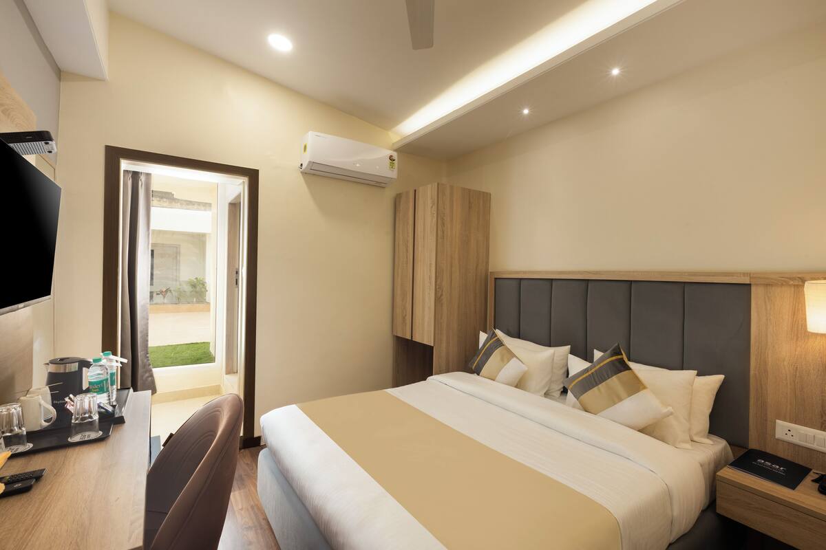 A hotel room with a bed, bedside table, and a desk with a chair. There is an air conditioning unit on the wall and a television on the desk.