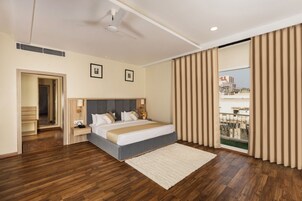 A modern hotel room with a large bed, wooden flooring, a ceiling fan, and a view of the city through floor-to-ceiling windows.