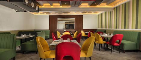 L’intérieur d’un restaurant présente des banquettes vertes, des chaises jaunes et rouges, et une table à manger centrale.