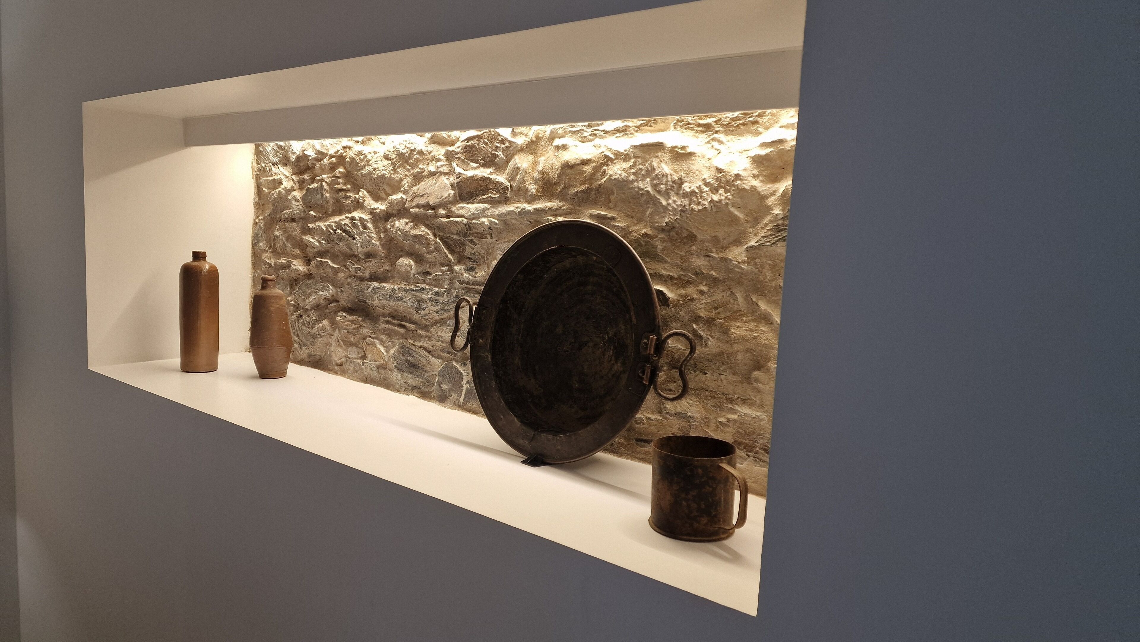 Eine Regalvorrichtung mit einer Steinwand im Hintergrund, mit einem großen schwarzen Teller, einer kleinen Vase und einer Metalltasse.