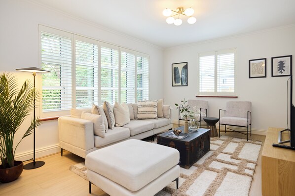 A modern living room with a sofa, ottoman, coffee table, and a chair. There are large windows with blinds, a floor lamp, and framed artwork on the wall. - The Lindens Garden Retreat (Taplow)