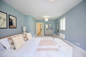 A bedroom with a large bed, bedside tables, a dresser, and a mirror. There is a door leading to another room. - The Lindens Garden Retreat (Taplow)