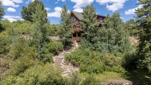 A wooden cabin with a staircase leading to a balcony, surrounded by dense forest.