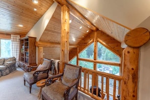 A log cabin interior with a high ceiling, wooden beams, a large window, and a fireplace.